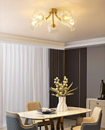 Load image into Gallery viewer, A sophisticated dining room illuminated by the Kalin Flower Glass Ceiling Light, featuring a brushed gold frame and delicate petal-shaped glass shades. The ceiling light&#39;s warm glow enhances the luxurious ambiance of the room, highlighting the elegant marble dining table, quilted chairs, and a striking centerpiece of dried flowers in a white ceramic vase. The floor-to-ceiling drapery adds a soft, refined touch to the modern decor.
