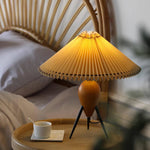 Load image into Gallery viewer, The Makie Table Lamp on a round bedside table, casting a warm glow next to a rattan headboard, creating a serene and inviting bedroom scene.
