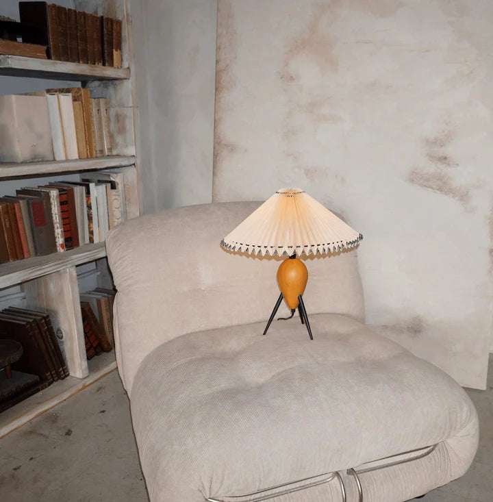 The Makie Table Lamp glowing warmly atop a soft beige tufted chair, styled next to a rustic bookshelf filled with classic books, creating an inviting reading nook ambiance.