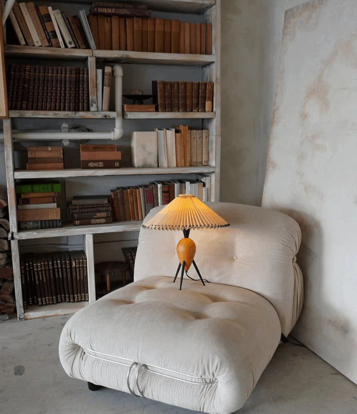 A cozy reading nook featuring the Makie Table Lamp with its pleated shade glowing warmly on a soft, tufted beige armchair. A rustic bookshelf filled with vintage books serves as a charming backdrop, enhancing the warm and inviting ambiance.