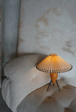 Load image into Gallery viewer, The Makie Table Lamp resting on a plush, tufted lounge chair, its warm pleated light casting a cozy glow against a distressed beige backdrop, creating a serene reading nook ambiance.
