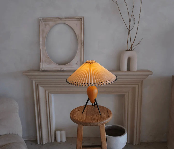 The Makie Table Lamp placed atop a rustic wooden stool in front of an elegant beige fireplace mantel. The pleated lamp shade emits a warm glow, complementing the minimalist decor featuring a round-frame art piece and modern ceramic vases.