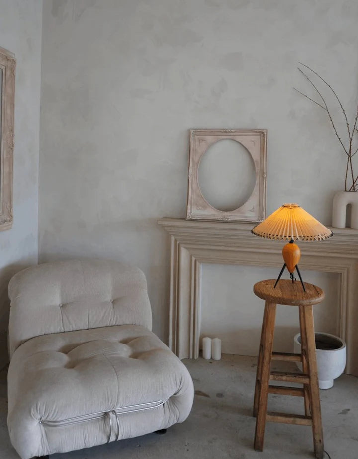 The Makie Table Lamp placed on a rustic wooden stool, illuminating a calming neutral corner with a tufted lounge chair, a minimal mantel, and decor accents that evoke understated elegance.