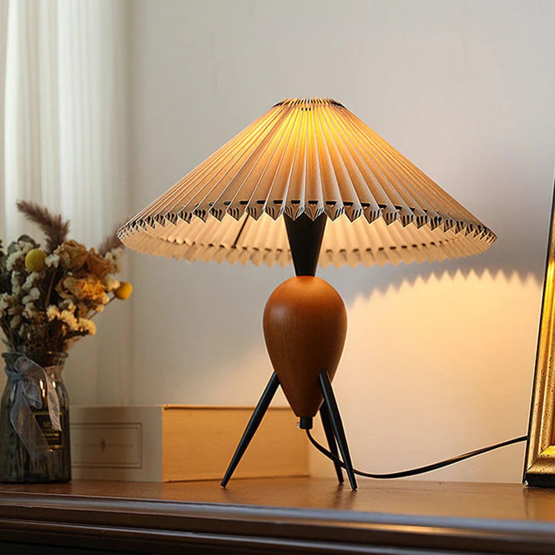 A cozy tabletop setup featuring the Makie Table Lamp with a unique pleated shade casting a warm ambient glow. The lamp's wooden, teardrop-shaped base is supported by sleek black tripod legs, creating an inviting and elegant aesthetic.