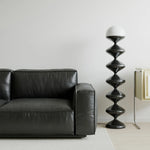 Load image into Gallery viewer, Minimalist home decor featuring a sleek Gourd Lamp with a spiral black base and a clean white top, placed next to a black leather sofa. The simple yet striking design complements the neutral, light-filled room, emphasizing a modern and understated elegance.
