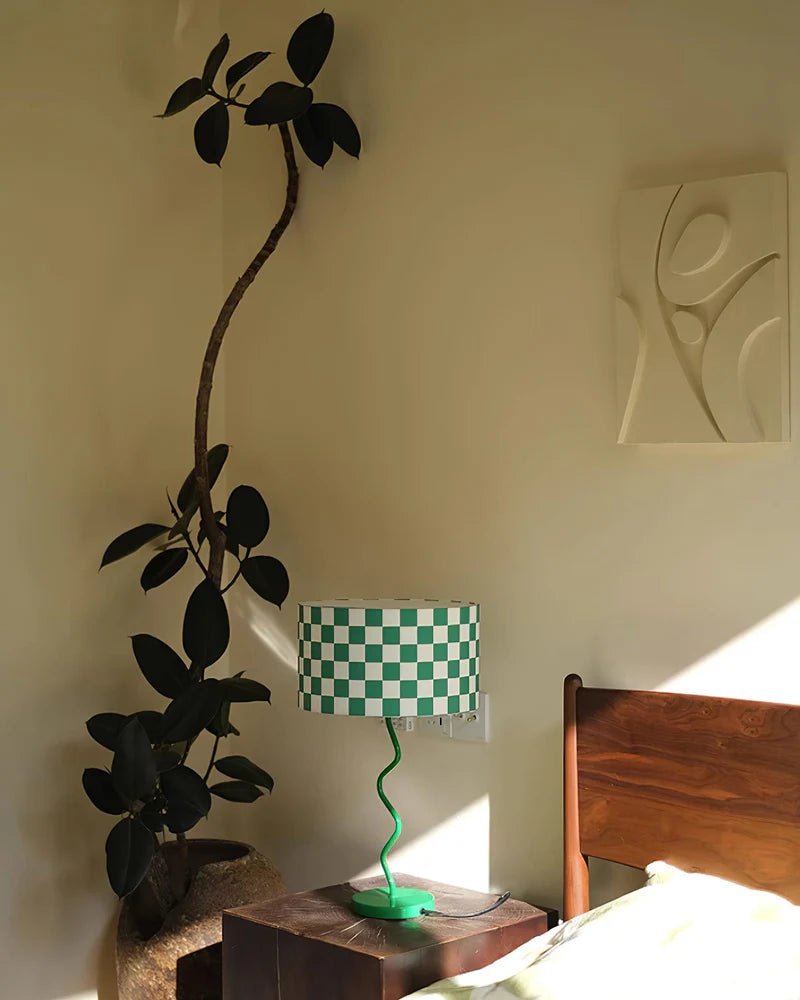 A modern bedroom corner featuring a Checker Lamp with a green and white checkered shade and a spiraled green base, placed on a wooden bedside table. The setting is enhanced by natural light streaming in, illuminating a tall indoor plant in the corner and a minimalist wall sculpture. The combination of organic elements and contemporary decor creates a serene and stylish ambiance.