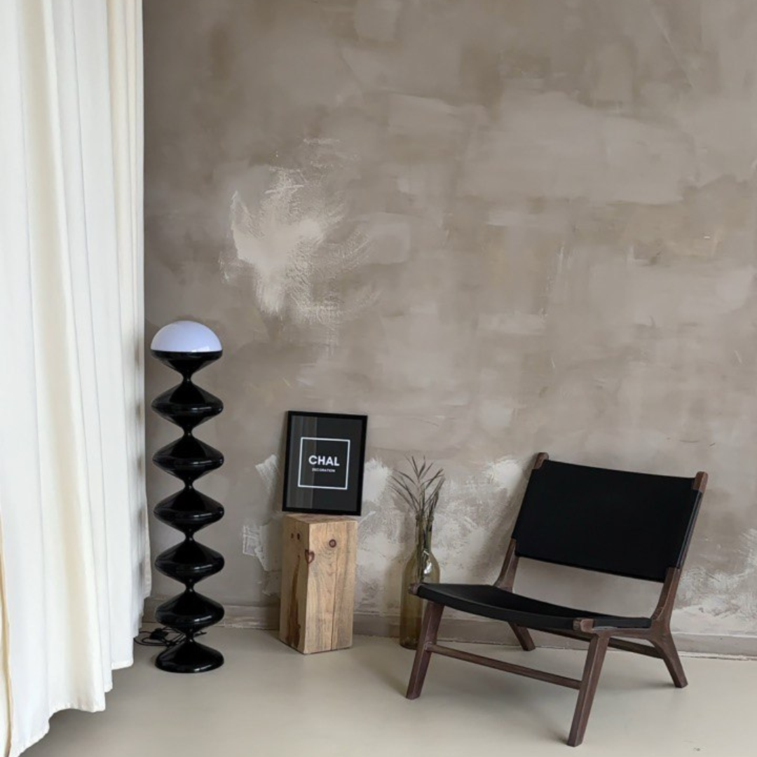 Contemporary room setting displaying a Modern Gourd Design Floor Lamp with stacked black gourd shapes and a white dome, placed beside a wooden block displaying a frame labeled 'CHAL', with a stylish black chair and a grey textured wall in the background.