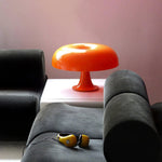 Load image into Gallery viewer, An orange Mushroom Playful Table Lamp shines brightly on a white table, contrasting vividly with the dark grey couch and complementing the soft pink wall, adding a splash of cheerful color to a stylish urban lounge space.
