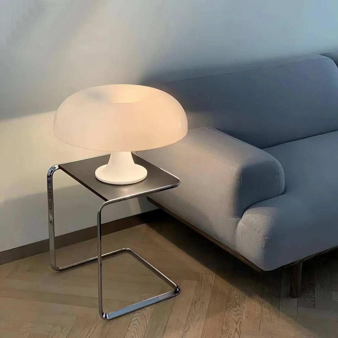 The Mushroom Playful Table Lamp in soft cream casts a warm, inviting glow on a sleek side table next to a contemporary grey sofa, adding a touch of modern elegance and cozy ambiance to the living space.