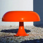 Load image into Gallery viewer, The vibrant orange Mushroom Playful Table Lamp stands out against a backdrop of white walls and pale gravel, its glossy finish reflecting the bright sunlight, adding a modern touch to an outdoor setting with its striking color and iconic shape.
