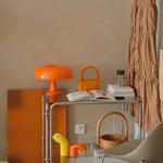 Load image into Gallery viewer, The Mushroom Playful Table Lamp in bright orange adds a pop of color to a stylishly curated corner, surrounded by harmoniously matching accessories like a chic bag and decorative items, creating a cohesive and vibrant interior aesthetic.
