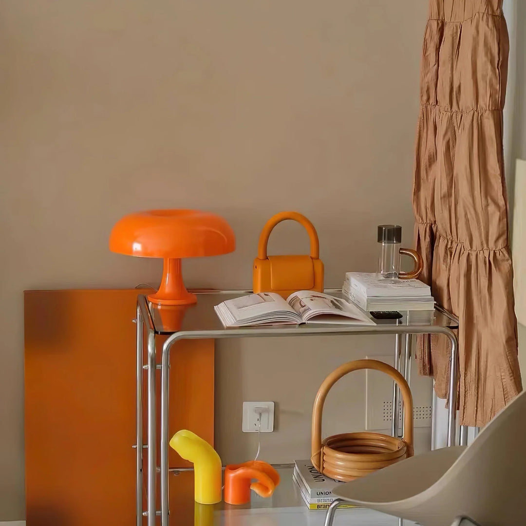 The Mushroom Playful Table Lamp in bright orange adds a pop of color to a stylishly curated corner, surrounded by harmoniously matching accessories like a chic bag and decorative items, creating a cohesive and vibrant interior aesthetic.