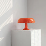 Load image into Gallery viewer, A striking orange Mushroom Playful Table Lamp stands out in a brightly sunlit corner, its smooth, reflective surface casting intriguing shadows against the minimalist white walls, enhancing the vivid and modern aesthetic of the space.
