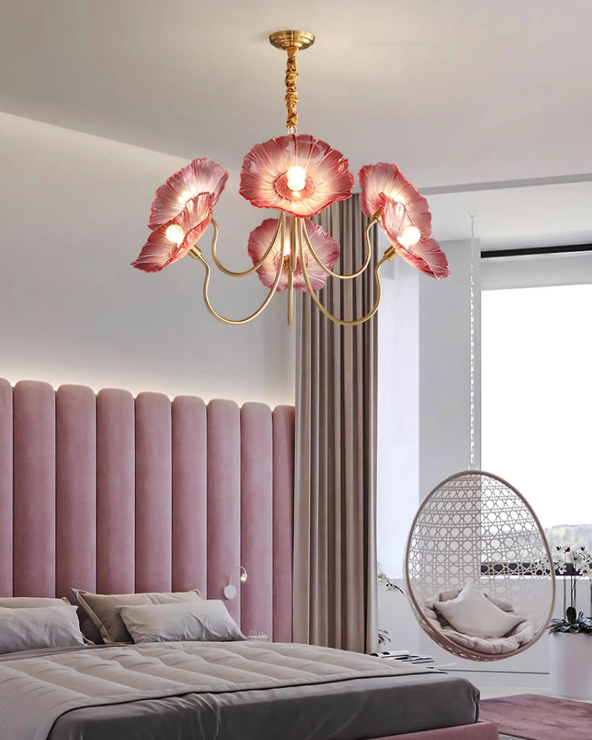 A luxurious bedroom illuminated by the Oleander Floral Chandelier featuring pink glass flower-like shades and a gold frame, hanging above a plush bed with a pink velvet tufted headboard, neutral bedding, and soft cushions. The room also features elegant drapery, a hanging rattan egg chair, and large windows allowing in natural light.