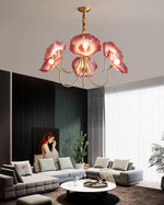 Load image into Gallery viewer, A captivating Oleander Floral Chandelier with vibrant pink glass petal shades and a gold frame hanging above a chic modern living room, featuring a gray sectional sofa with white and pastel cushions, a round marble coffee table, and bold accent decor, including a vertical slat panel wall and contemporary artwork.
