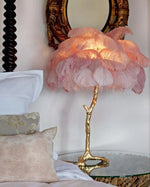 Load image into Gallery viewer, Pink feather palm table lamp with a golden branch base placed on a glass bedside table, styled in a cozy bedroom with ornate vintage mirror and soft decorative pillows.
