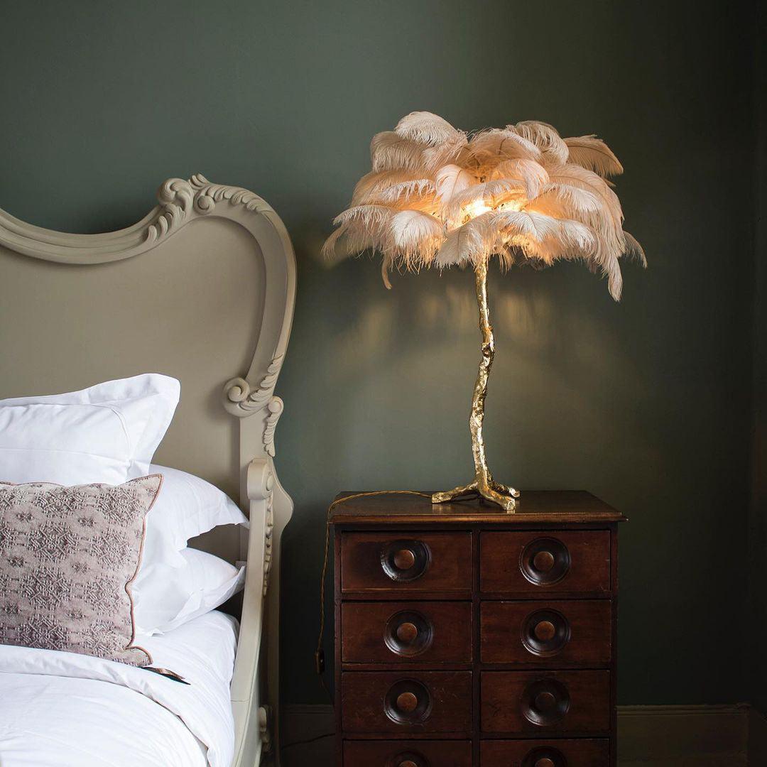 Neutral-toned feather palm table lamp with a gold branch stand, placed on a vintage dark wood bedside cabinet, creating a cozy and elegant ambiance in a bedroom with classic decor.