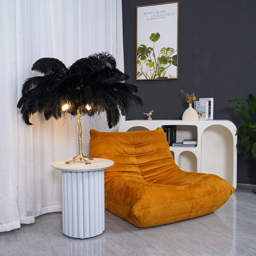 Black feather palm table lamp with a gold tree-inspired base placed on a white cylindrical side table next to a mustard yellow lounge chair, styled with a modern white bookshelf and potted greenery in a cozy room.