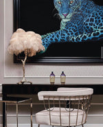 Load image into Gallery viewer, Cream feather palm table lamp with a gold tree-like base placed on a sleek black console table, styled with a dramatic blue panther artwork in the background and complemented by a modern silver-framed chair.
