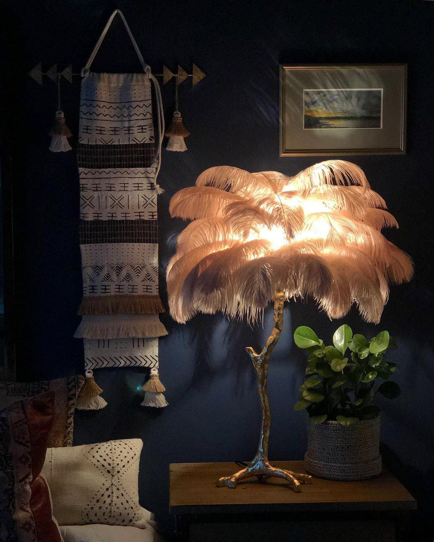 Feather palm table lamp with a gold branch base casting a warm glow in a bohemian-style room featuring dark navy walls, tribal wall decor, and a potted plant for added texture and ambiance.