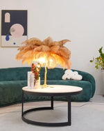 Load image into Gallery viewer, Gold feather palm table lamp with a sculpted tree trunk base placed on a round modern coffee table, styled with a green velvet couch, abstract wall art, and decorative accents in a chic living room.
