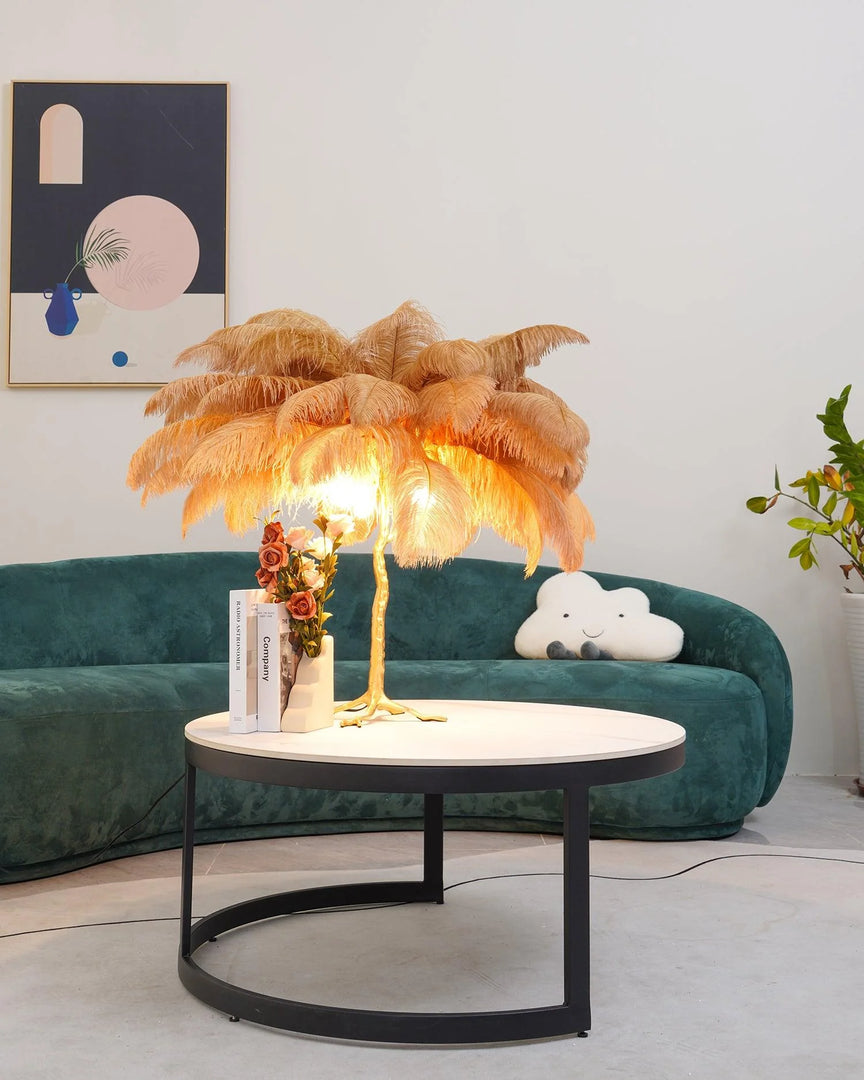 Gold feather palm table lamp with a sculpted tree trunk base placed on a round modern coffee table, styled with a green velvet couch, abstract wall art, and decorative accents in a chic living room.