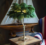 Load image into Gallery viewer, Green feather palm table lamp with a golden branch base placed on a wooden side table, styled in a nautical-themed room with a decorative ship model and a navy-striped accent chair.
