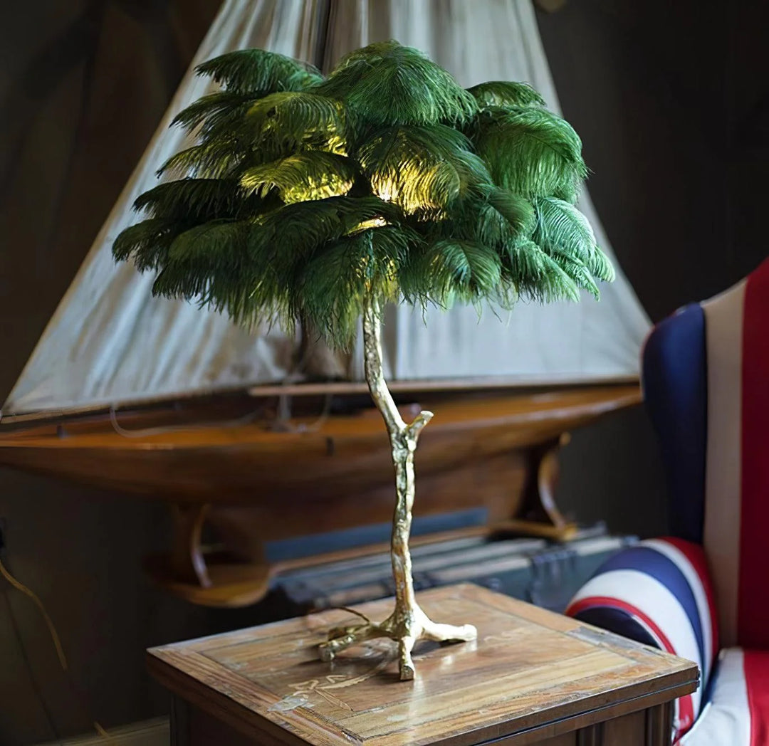 Green feather palm table lamp with a golden branch base placed on a wooden side table, styled in a nautical-themed room with a decorative ship model and a navy-striped accent chair.