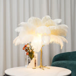 Load image into Gallery viewer, White feather palm table lamp with a gold tree-inspired base placed on a round marble table, styled with a modern floral arrangement and a velvet green armchair in the background.
