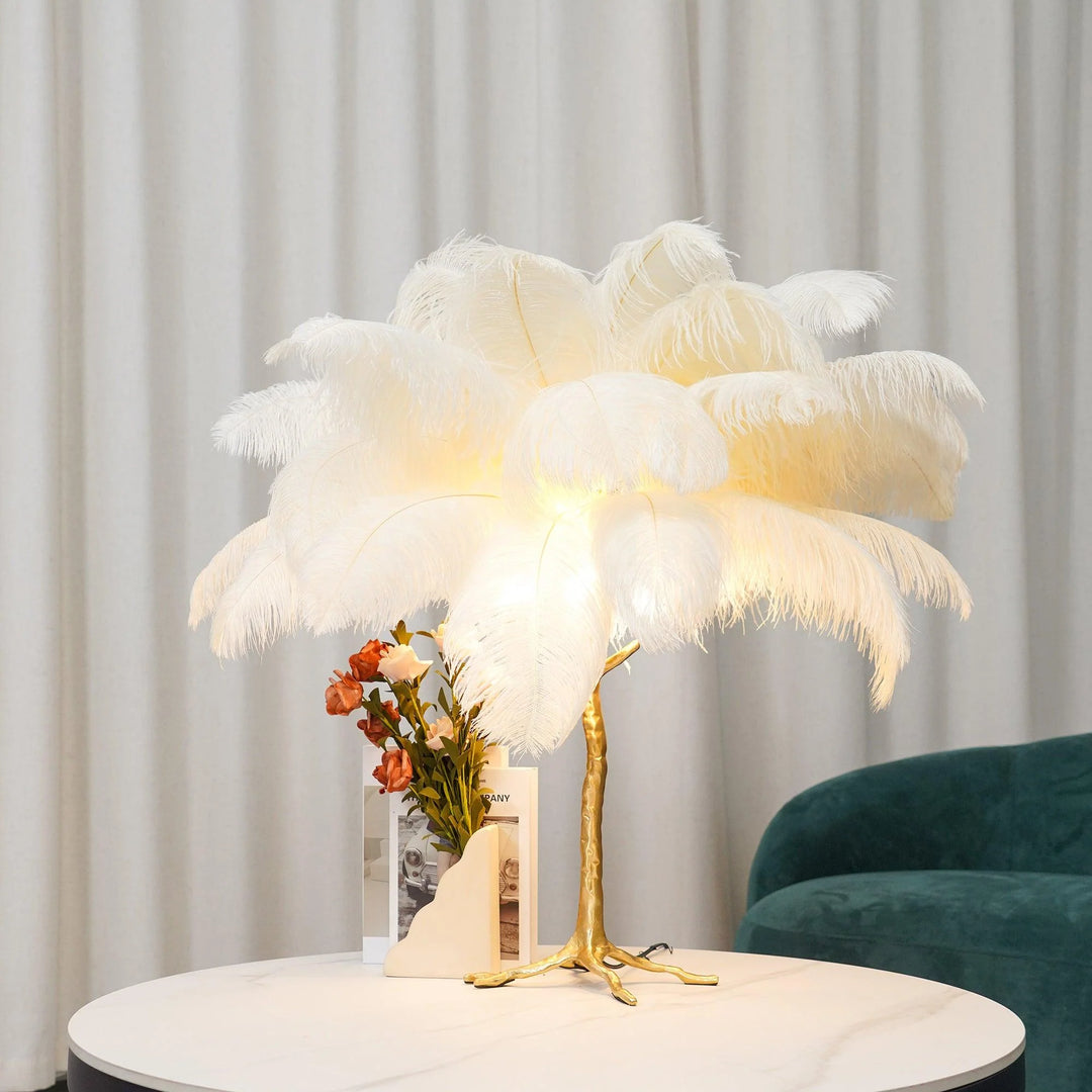 White feather palm table lamp with a gold tree-inspired base placed on a round marble table, styled with a modern floral arrangement and a velvet green armchair in the background.