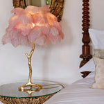 Load image into Gallery viewer, Elegant pink feather palm table lamp with a gold branch-like stand placed on a glass-topped bedside table, adding a luxurious and whimsical touch to a classic bedroom setting.
