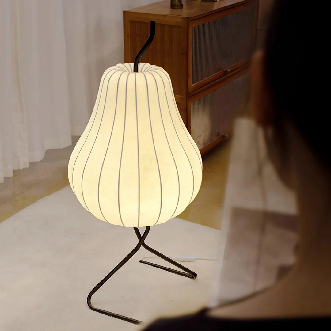 Close-up view of a glowing pear-shaped floor lamp with vertical stripes and a black tripod stand, placed on a soft white rug near a wooden sideboard in a cozy living space.