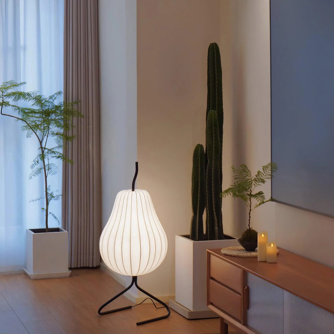 Softly glowing pear-shaped floor lamp with a black tripod stand, placed in a modern living room corner near a cactus and potted fern, complemented by candles and a wooden sideboard.