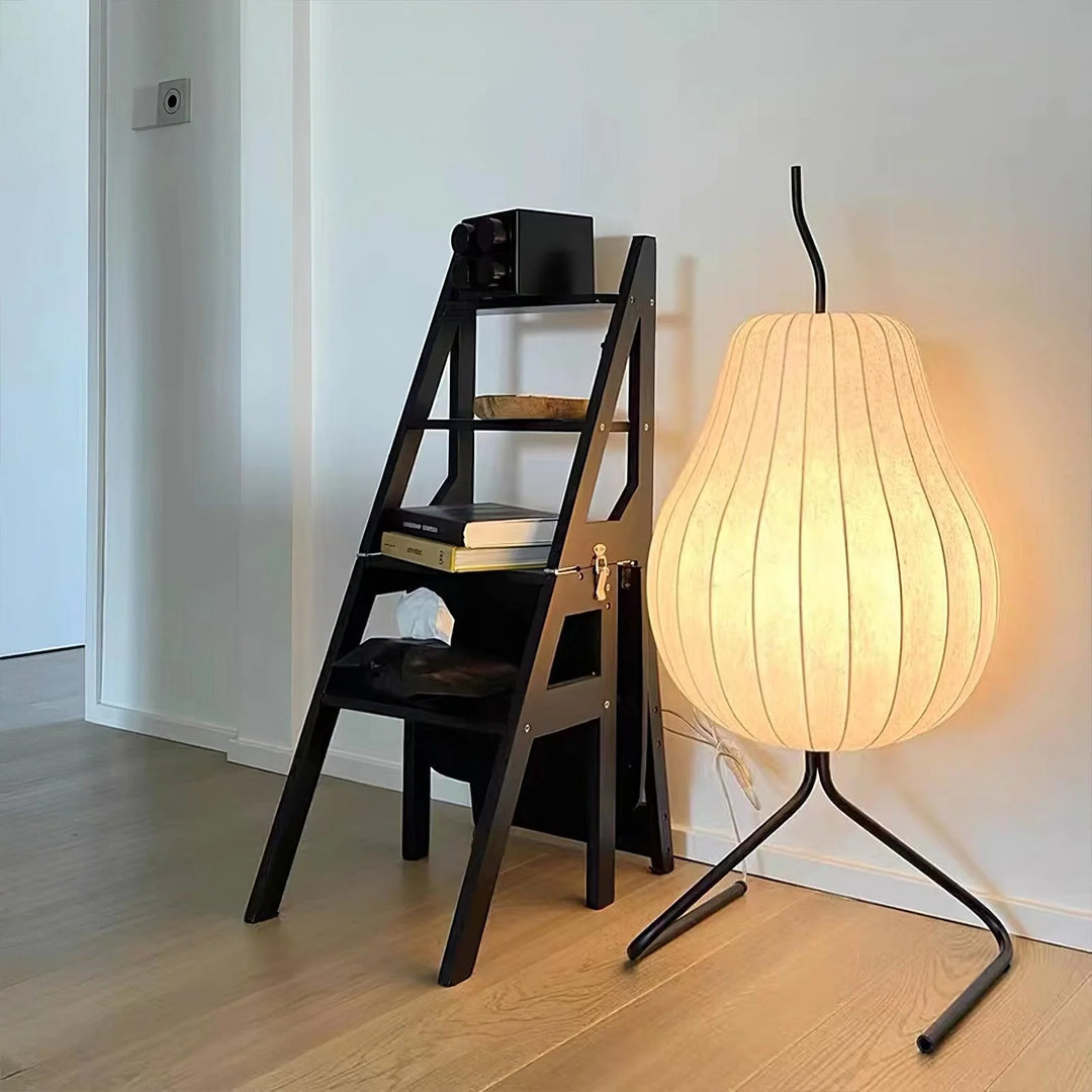 Modern pear-shaped floor lamp with a glowing cream shade and a black metal stand, placed beside a minimalist black step-shelf in a bright room with wooden flooring.