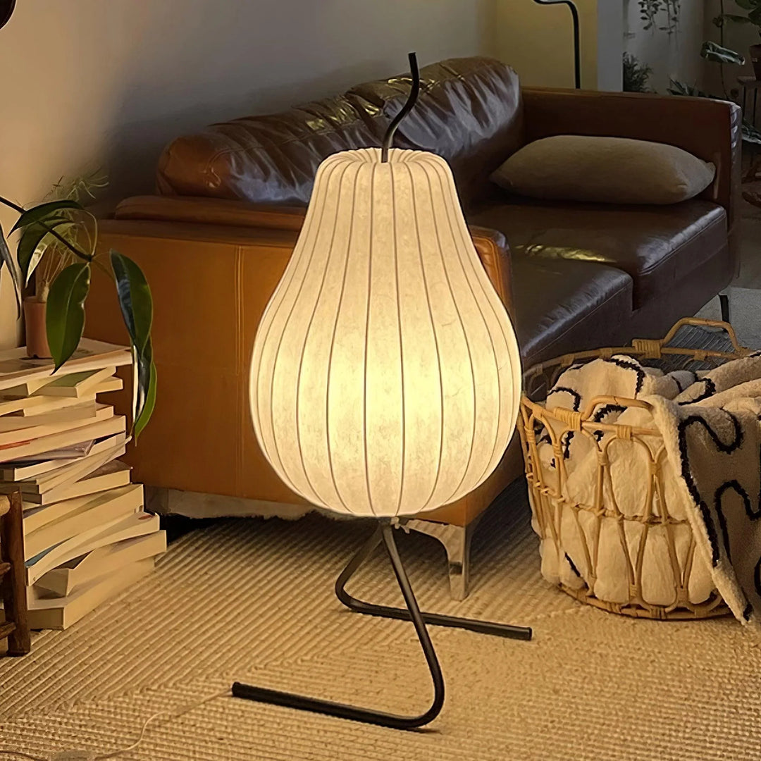 Elegant pear-shaped floor lamp with a soft glowing shade and black curved metal stand, placed in a cozy living room next to a leather couch, basket, and stacked books.