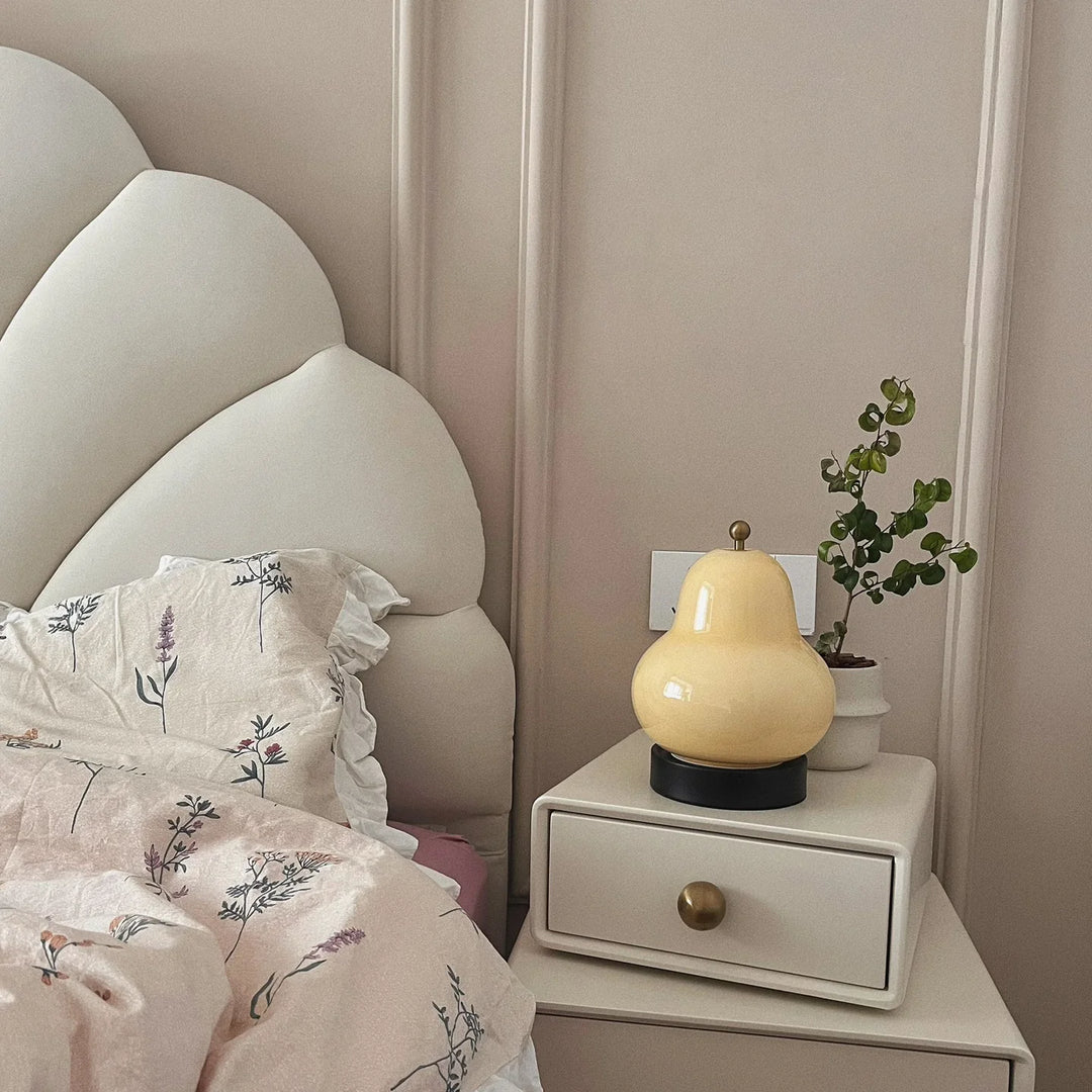 A pear-shaped bedside lamp with a warm glow sits elegantly on a white nightstand, accompanied by a potted plant, soft floral bedding, and a tufted headboard for a cozy bedroom setup.