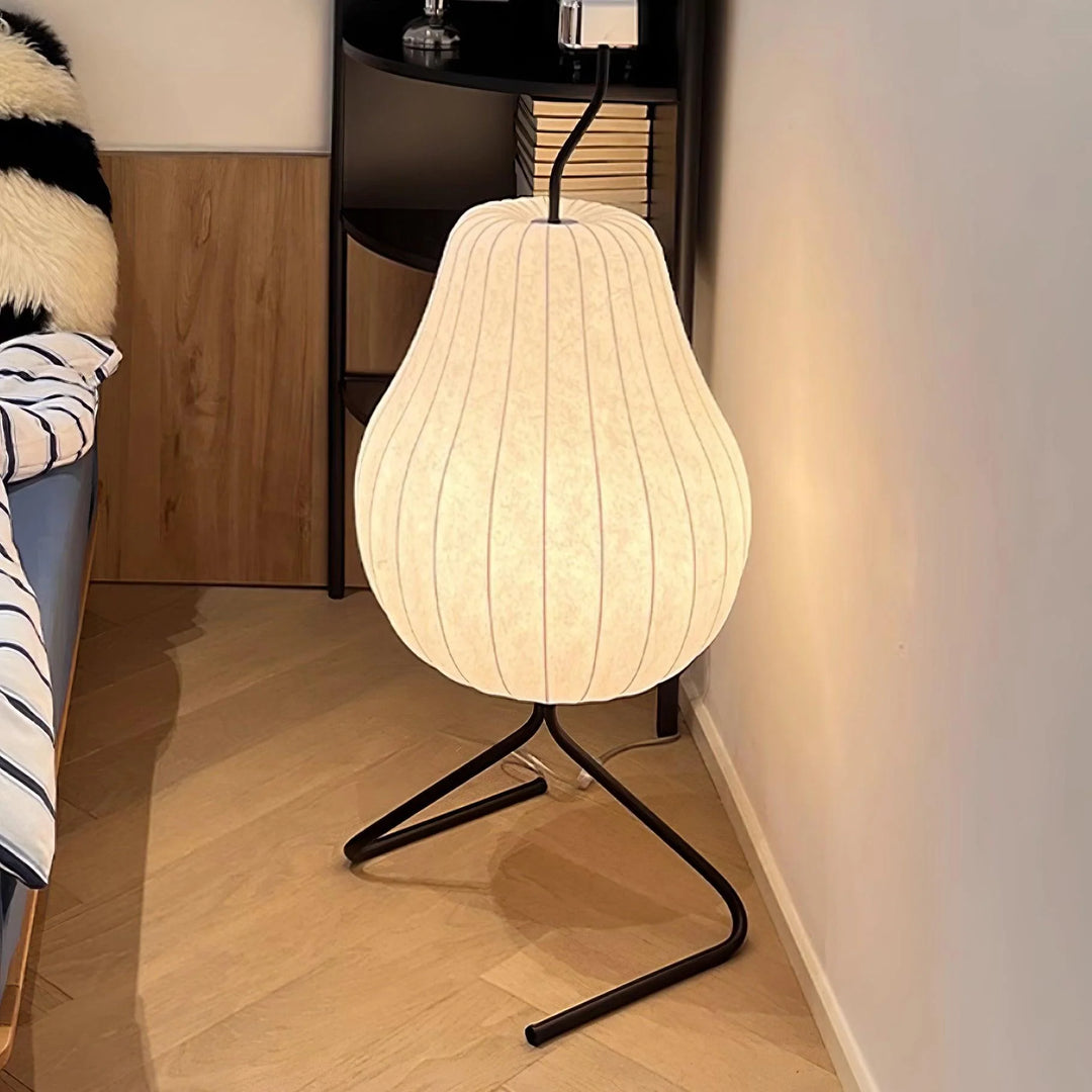 Minimalist pear-shaped floor lamp with a soft glowing white shade and black frame, placed beside a cozy bed and a modern bookshelf.