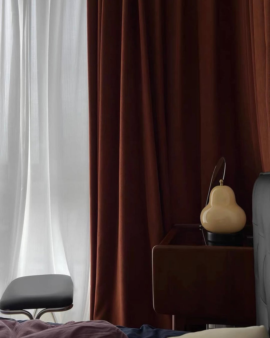 A close-up view of a glowing pear-shaped lamp sitting on a dark wooden dresser, set against rich burgundy curtains and sheer white drapes, adding an ambient touch to the cozy room decor.