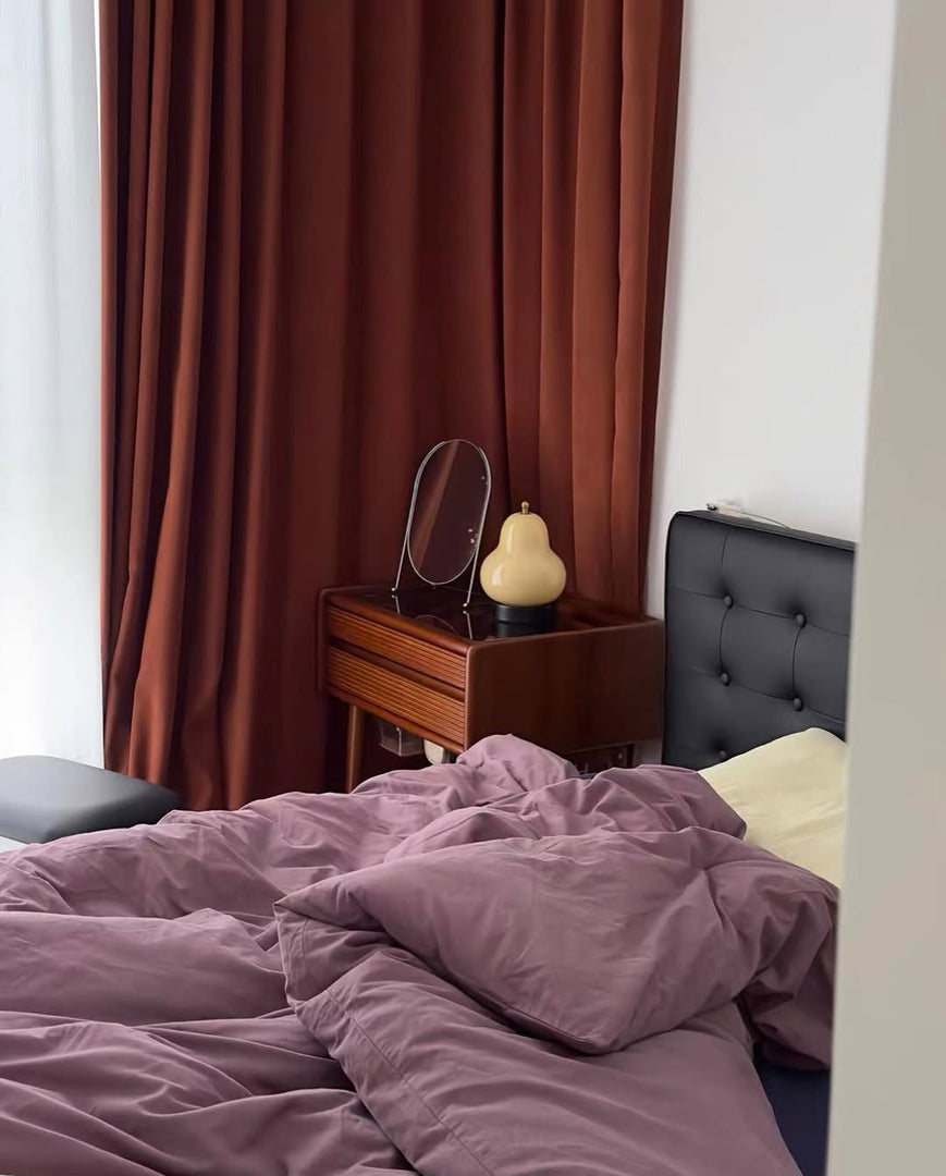 A warm-glowing pear-shaped lamp placed on a mid-century modern wooden dresser next to a round mirror, complementing dark red curtains and a tufted black bedframe in a cozy, contemporary bedroom setting.