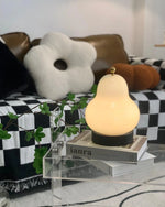 Load image into Gallery viewer, Illuminated pear-shaped table lamp resting on a clear acrylic stand with stacked books, complementing a cozy living room setup featuring checkered throws and unique decorative pillows.
