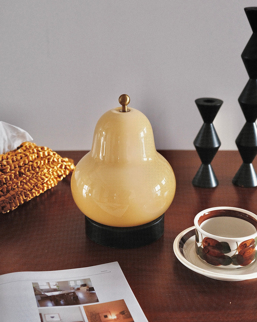 A close-up view of a pear-shaped table lamp placed on a wooden surface, styled with a vintage tea set, a decorative geometric ornament, and an open book, adding a cozy and refined aesthetic to the setup.