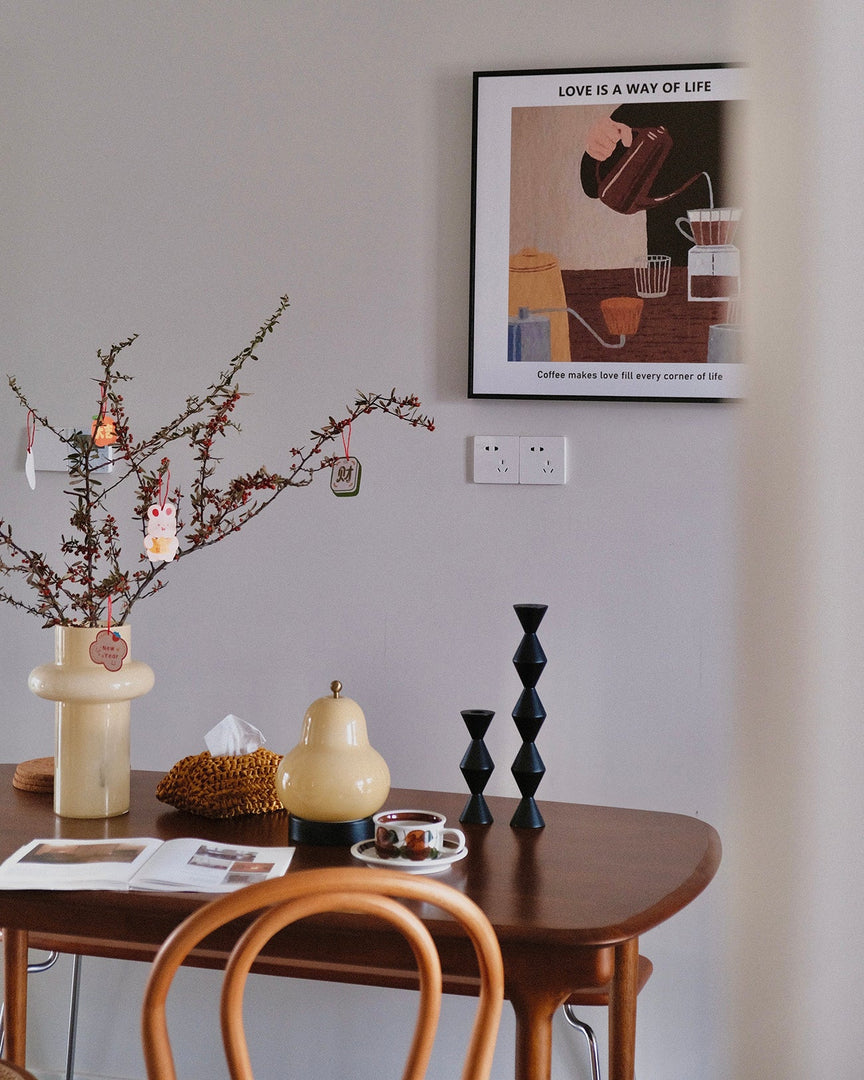 A pear-shaped table lamp placed on a wooden dining table alongside decor elements such as a branch arrangement in a vase, geometric candlesticks, and a coffee set, creating a cozy and artistic dining space.