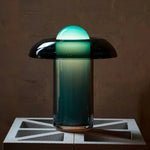 Load image into Gallery viewer, Modern Persian green table lamp with a glowing dome-shaped top and a gradient glass base, placed on a geometric concrete block against a textured wall.
