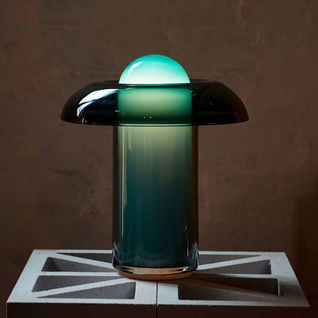 Modern Persian green table lamp with a glowing dome-shaped top and a gradient glass base, placed on a geometric concrete block against a textured wall.