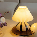 Load image into Gallery viewer, Pleated Cavolo table lamp with a glowing pleated shade and striped ceramic base, placed on a round tinted tray alongside floral decor in a modern living space.
