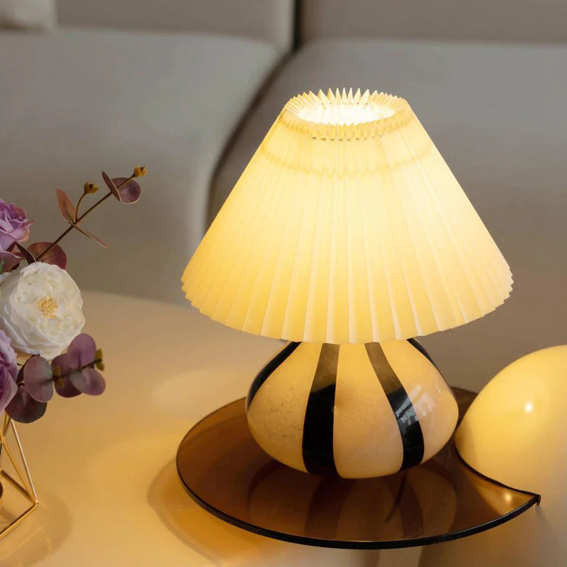 Pleated Cavolo table lamp with a glowing pleated shade and striped ceramic base, placed on a round tinted tray alongside floral decor in a modern living space.