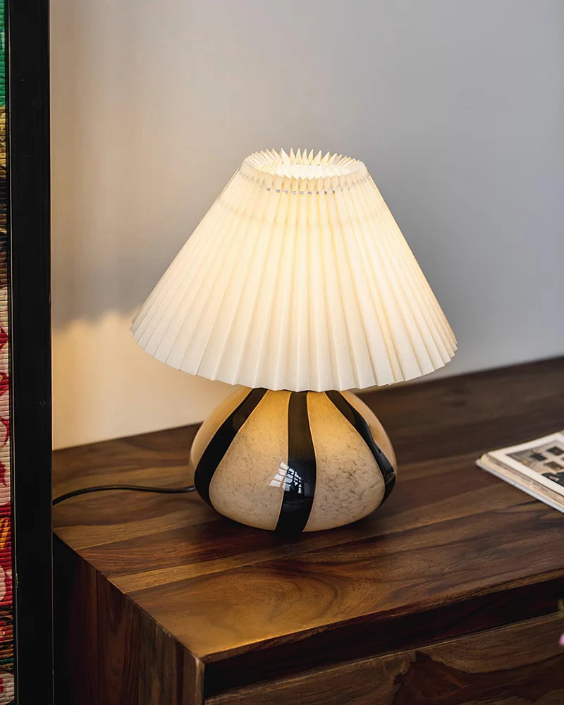 Pleated Cavolo table lamp with a striped ceramic base and soft glowing pleated shade, placed on a dark wooden dresser beside neatly arranged reading materials.