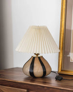 Load image into Gallery viewer, Pleated Cavolo table lamp with a striped ceramic base and a cream pleated shade, placed on a wooden surface next to a gold-framed artwork for an elegant decor accent.
