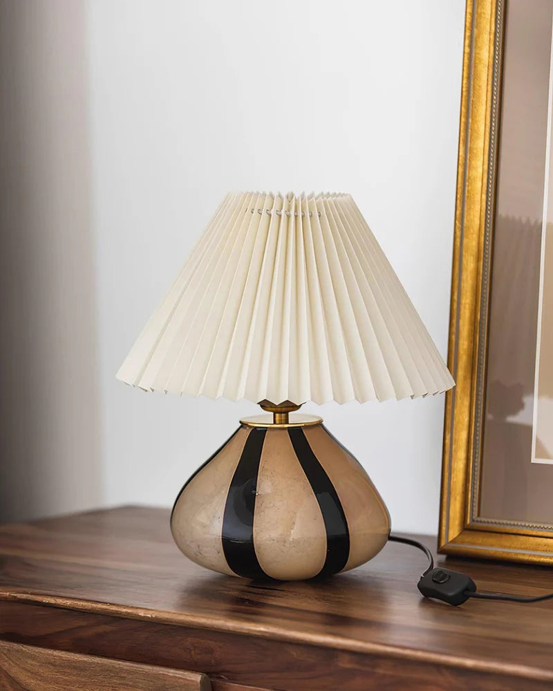 Pleated Cavolo table lamp with a striped ceramic base and a cream pleated shade, placed on a wooden surface next to a gold-framed artwork for an elegant decor accent.