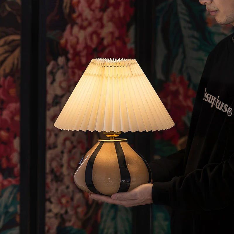 Pleated Cavolo table lamp with a glowing pleated shade and striped ceramic base, held by a person in front of a vibrant floral backdrop, highlighting its elegant design and soft light.